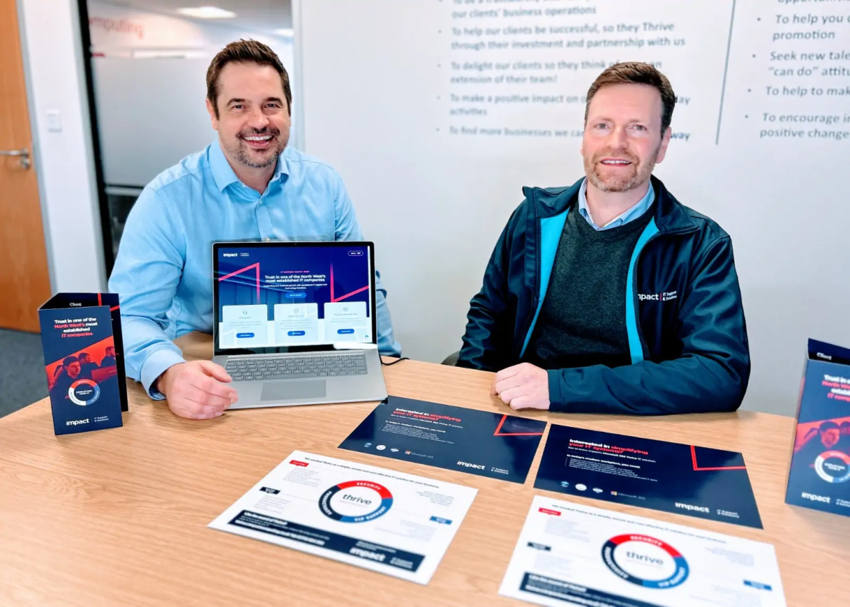 Adam Gregson from Impact IT Support & Solutions sitting with Simon Cocker at Voodoo Agency at a table with a laptop showing the Impact website, surrounded by branded marketing materials including 'Thrive' IT solution leaflets and folders.